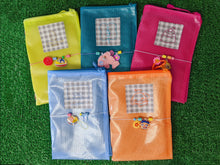 Load image into Gallery viewer, five colorful mesh zippered pouch journal covers arranged on green artificial grass. the pouches are yellow, teal, pink, light blue, and orange, each with a gingham patch containing unique embroidered designs and small decorative charms.