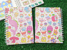 Load image into Gallery viewer, two spiral-bound notebooks showing the front and back of a colorful cartoon character pattern, placed on a green grass-like surface. in the background, there is an open notebook showing the sticker-collecting wax paper inside.