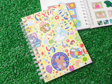 Load image into Gallery viewer, one spiral-bound notebooks with a colorful stickerbomb inspired pattern, placed on a green grass-like surface. behind it, there is an open notebook showing the sticker-collecting wax paper inside.