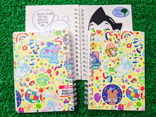 Load image into Gallery viewer, two spiral-bound notebooks showing the front and back of a colorful stickerbomb inspired pattern, placed on a green grass-like surface. in the background, there is an open notebook showing the sticker-collecting wax paper inside.