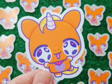 Load image into Gallery viewer, a close-up of a hand holding a die-cut sticker of an orange cartoon unicorn character with purple and yellow details, set against a background of multiple identical stickers on a green textured surface.
